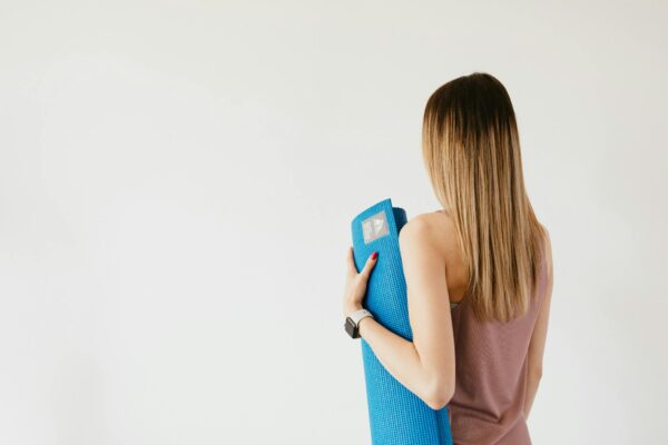 Back view of unrecognizable female in activewear standing near gray wall and holding yoga mat in hand